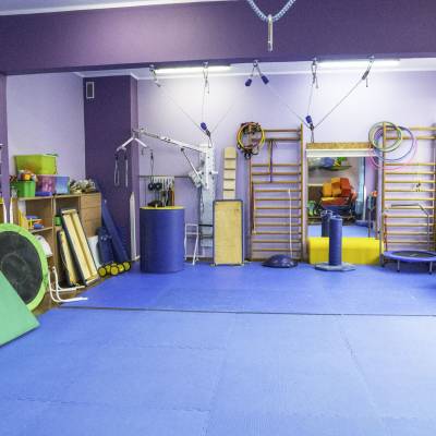Sala terapii SI: niebieskie maty, drabinki, lustro i kolorowe przybory do ćwiczeń przy oknach.